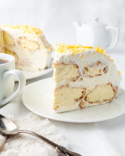 Um pedaço de bolo de creme com chantilly Decorado com gotas de chocolate amarelas Feito em casa em um prato branco Perto de uma colher e uma caneca No fundo está um prato com bolo e um bule de chá branco