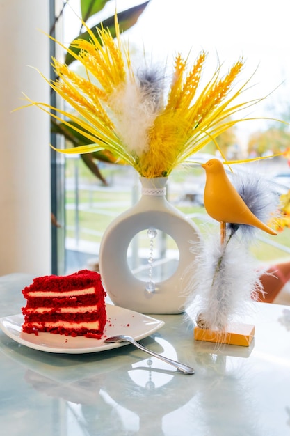 Um pedaço de bolo delicioso em um prato e uma xícara de café na mesa Há flores amarelas artificiais e um pássaro de brinquedo na mesa do restaurante Mesa de centro perto da janela