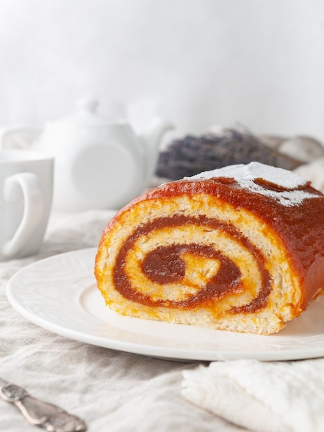 Um pedaço de pão-de-ló caseiro com geleia de maçã em um prato branco no fundo está um bule de chá branco e uma caneca.