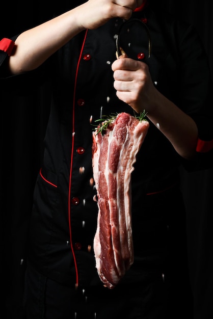 Foto um pedaço de porco fresco em um gancho de carne nas mãos de um chef carne em um fundo preto bacon