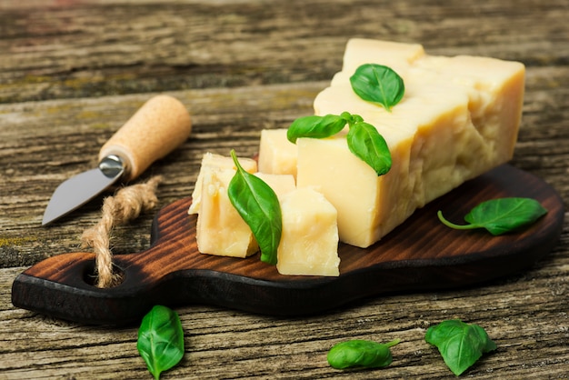 um pedaço de queijo parmesão com folhas de manjericão e faca de queijo em uma placa de madeira