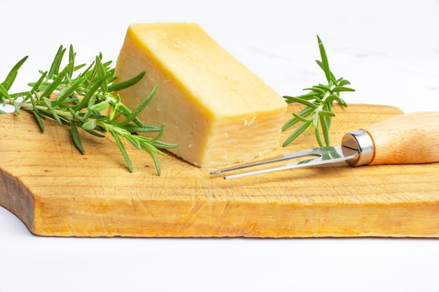 Um pedaço de queijo parmesão um garfo e galhos de alecrim em uma placa de cozinha de madeira