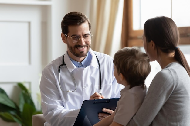 Um pediatra sorridente de uniforme médico branco segurando um caderno ouvindo uma jovem mãe com um filho em uma reunião de check-up. Conceito de cuidados de saúde de consulta médica profissional para crianças