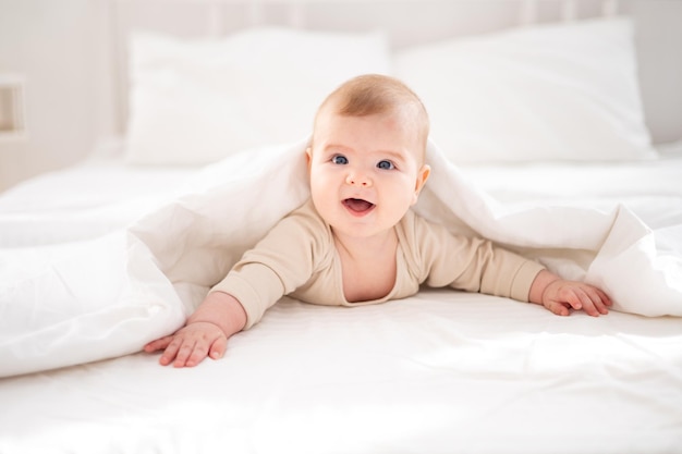 Um pequeno bebê saudável em uma roupa de algodão está de bruços debaixo de um cobertor em uma cama na cama branca no quarto em casa olha para a câmera sorri