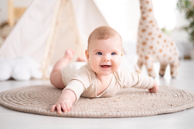 Um pequeno bebê saudável em uma roupa de algodão está de bruços em um tapete na sala de estar da casa no contexto de uma tenda e brinquedos de pelúcia