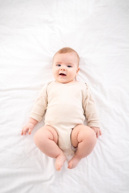 Um pequeno bebê saudável em uma roupa de algodão está deitado de costas em uma cama na cama branca no quarto em casa olha para a câmera sorri