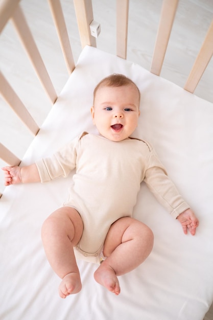 Um pequeno bebê saudável em uma roupa de algodão está deitado de costas na cama de uma criança na cama branca no quarto da casa olha para a câmera sorri