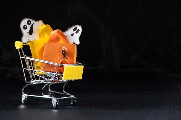 Um pequeno carrinho com sacos de presentes e fantasmas no fundo preto Halloween Venda espaço de cópia para texto