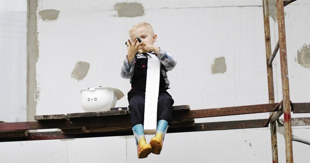 Um pequeno construtor com um capacete branco está sentado no andaime com um alicate nas mãos