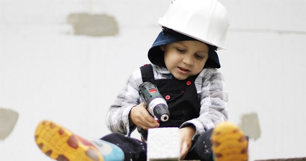 Um pequeno construtor de capacete branco está sentado no andaime com um alicate e uma broca ou chave de fenda