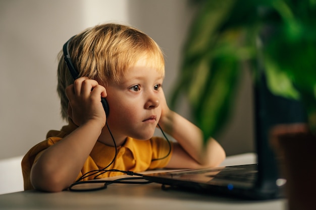 Foto um pequeno estudante interessado com fones de ouvido está sentado em sua mesa de casa e assistindo a um vídeo tutorial