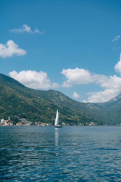 Um pequeno iate à vela flutua no mar pelas montanhas verdes, céu azul e nuvens