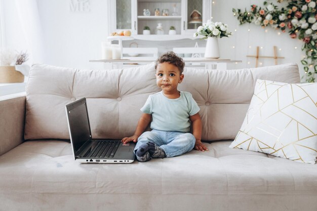 Um pequeno menino de pele escura afro-americano senta-se em um sofá com um laptop