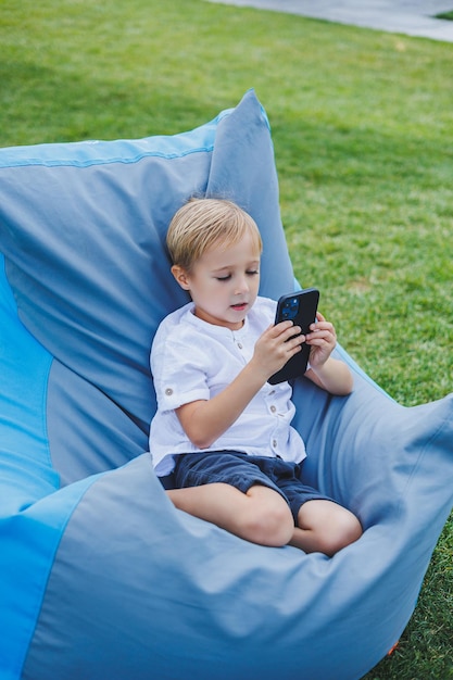 Um pequeno menino louro de 5 anos senta-se em uma cadeira na rua e olha para o telefone Uma criança moderna com um gadget