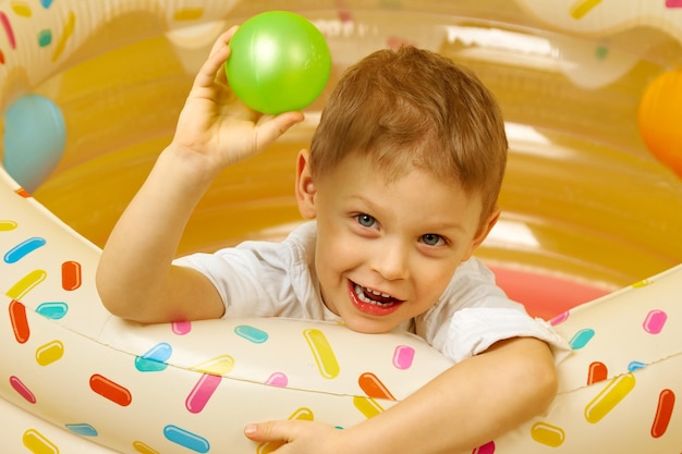 Um pequeno menino sorridente joga em uma piscina seca em um fundo amarelo. Uma criança joga uma bola de plástico verde