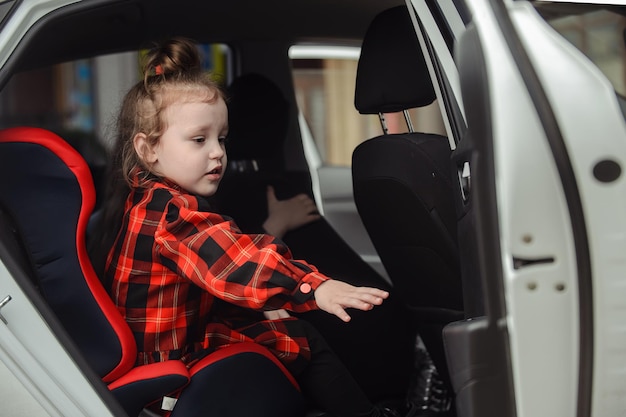 Foto um pequeno pré-escolar senta-se no banco de trás de um carro em uma cadeira de criança e tenta fechar a porta do carro
