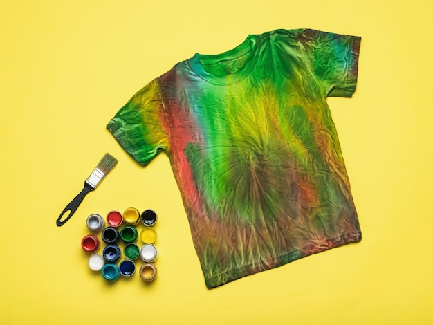 Um pincel, uma grande quantidade de tintas para tecidos e uma camiseta tie dye sobre fundo amarelo. Postura plana.