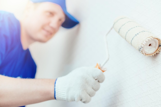 Um pintor de boné e luvas pinta a parede com um rolo de pintura Retrato de um jovem trabalhador de uniforme