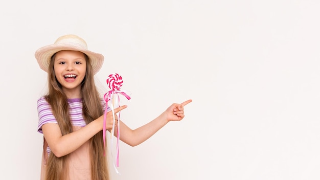 Um pirulito de caramelo nas mãos de uma linda garotinha Um bebê em um vestido de verão e um chapéu de verão aponta para um fundo branco isolado com o dedo indicador