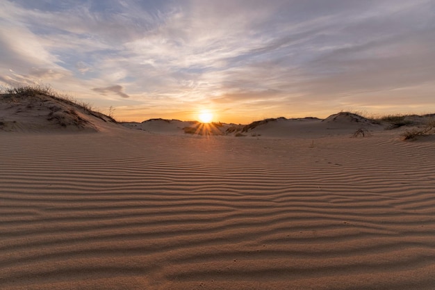 um pôr-do-sol com dunas de areia e o sol a pôr-se atrás delas