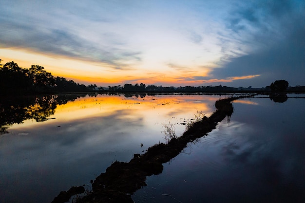 Um pôr do sol sobre um campo inundado com um rio em primeiro plano e um céu nublado ao fundo.