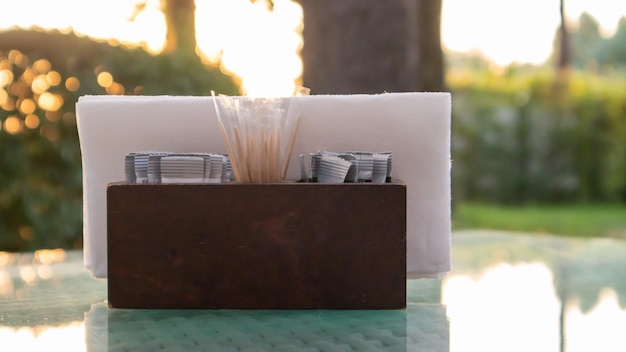 Um porta-guardanapos de madeira marrom e palitos de dente estão sobre a mesa de vidro do café, iluminada pelos raios do sol. configuração de mesa no restaurante. mesa de vidro em um jardim verde ao ar livre.