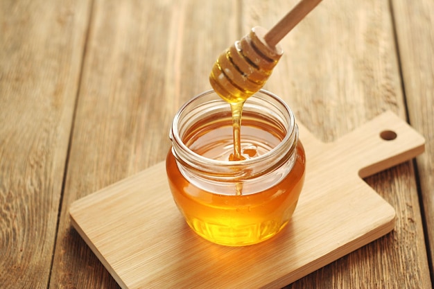 Um pote transparente de mel fica em uma mesa de madeira O mel flui para um pote em um palito de mel