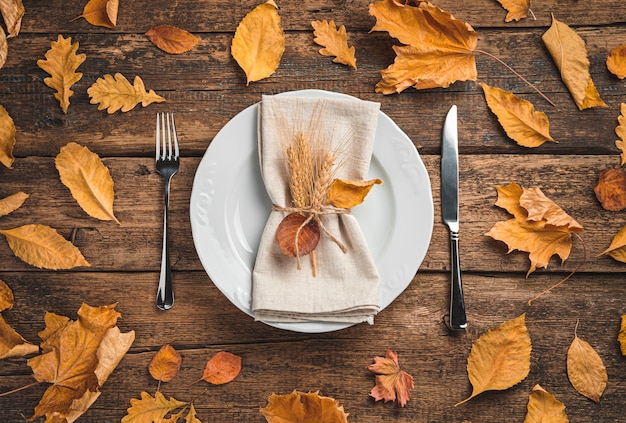 Um prato branco com um guardanapo e talheres em um fundo de madeira com folhas de outono