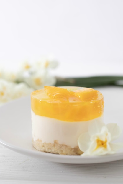 Um prato branco com um pedaço de cheesecake de manga sobre um fundo branco com uma flor ao fundo.