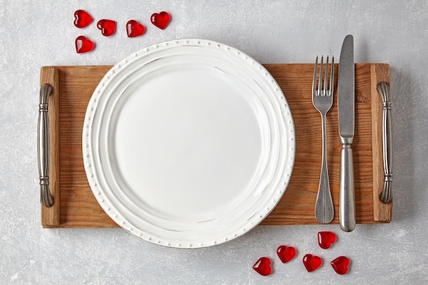 Um prato branco vazio com talheres em uma bandeja de madeira e corações de caramelo vermelho em uma mesa de concreto leve