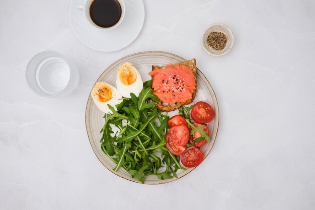 Um prato com um sanduíche de salmão rúcula tomate ovo cozido com copo de café e copo