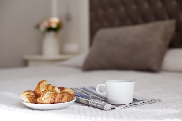 Um prato de croissants frescos, uma xícara de café e um jornal matinal na cama no quarto