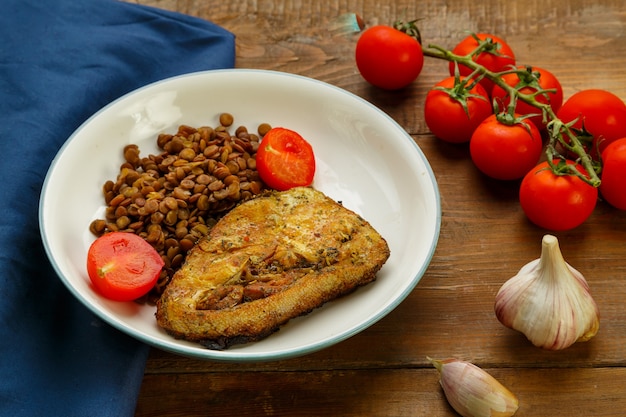 Um prato de peixe com lentilhas e tomates em uma mesa redonda com um garfo em um guardanapo azul ao lado de tomates cereja e limão.