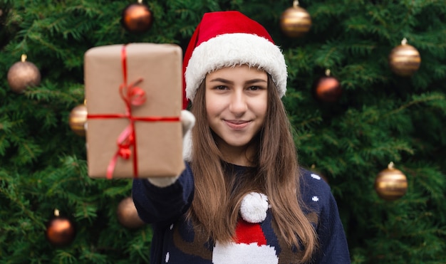 Um presente de Natal. Uma jovem com um chapéu de Papai Noel dá um presente feito de papel artesanal com uma fita vermelha. Contra o fundo de uma árvore de Natal