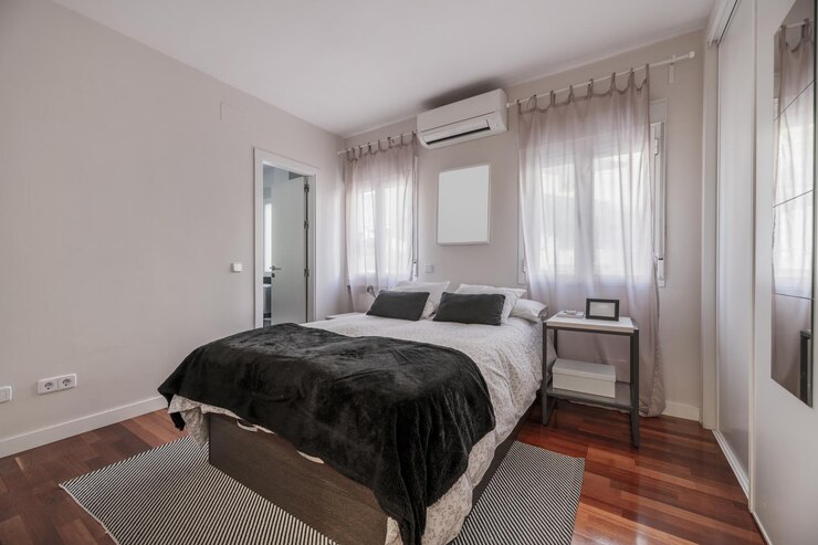 Foto um quarto com uma cama dupla com um armário embutido com portas deslizantes de madeira branca cobrindo uma parede e um ar condicionado acima da cama e uma porta de acesso a um banheiro