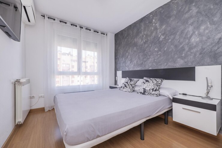 Foto um quarto duplo com uma cama grande sem edredão com mesas de cabeceira cantilever cabeceira preta e branca janela com cortinas brancas semitransparentes e radiador de alumínio