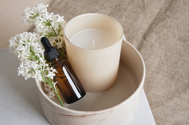 Um ramo de lilás branco, vela de aroma em um vidro e garrafas marrons com óleo de aroma em uma tigela de cerâmica em um fundo de linho natural.
