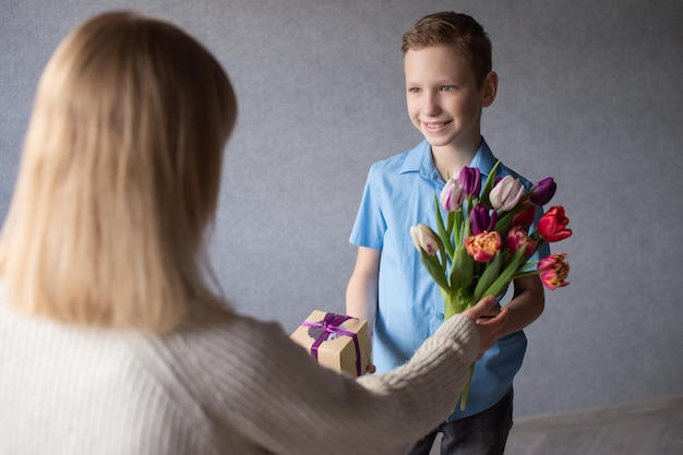 Um rapaz bonito a sorrir e a olhar para a mãe a dar flores e presentes para o Dia da Mãe.