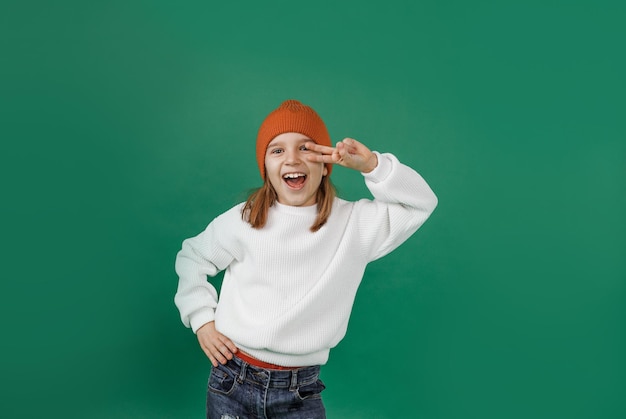 Um rapaz bonito com um suéter branco e um chapéu sorri e dança segurando os dedos perto dos olhos sobre um fundo verde isolado.