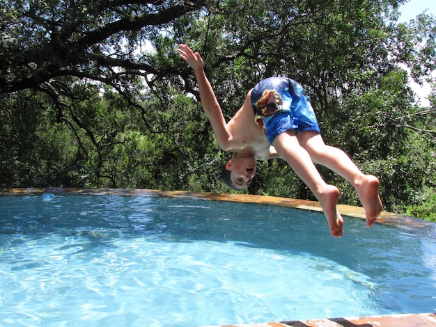 Foto um rapaz sem camisa a saltar para a piscina contra as árvores.