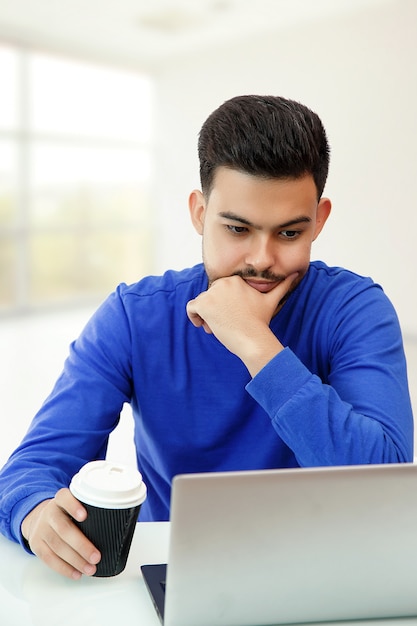 Um rapaz sentado em um laptop em busca de trabalho na Internet, fazendo negócios na rede global com uma xícara de café. Na luz