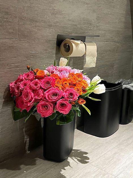Um recipiente preto com flores fica em um banheiro com papel higiênico na parede.