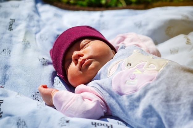 Um retrato da foto de um bebê com um boné vermelho encontra-se sobre uma manta branca na grama verde. A primavera está na rua, o sol está brilhando na criança