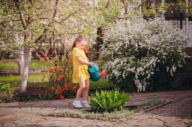 Um retrato de corpo inteiro de uma menina em um vestido amarelo regando flores de um regador de jardim azul.