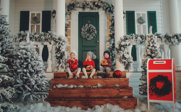 Foto um retrato de crianças sentadas na varanda de sua casa juntas, comer bagels. feliz natal feliz ano novo. quintal com uma árvore de natal, luzes e enfeites. hora do milagre.