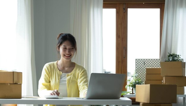 Um retrato de funcionário de comércio eletrônico de mulher asiática sentado no escritório cheio de pacotes na mesa usando um laptop e calculadora para tecnologia de comércio eletrônico de negócios de PME e negócios de entrega