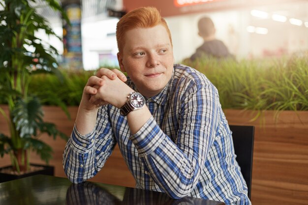 Um retrato de homem ruivo com sardas, vestindo camisa xadrez elegante e assistir sentado no café acolhedor, à espera de seus amigos