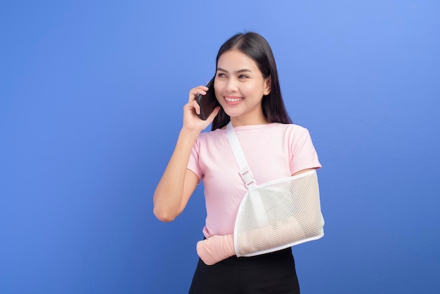 Um retrato de jovem com um braço ferido em um estilingue usando um telefone inteligente sobre fundo azul no conceito de estúdio, seguro e saúde