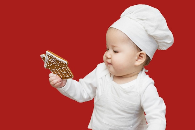 Um retrato de um bebê asiático em um chapéu de chef e avental olhando para um biscoito de gengibre de Natal isolado em um fundo vínico