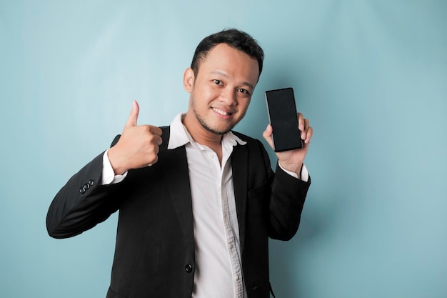 Um retrato de um empresário asiático feliz está sorrindo e segurando seu smartphone mostrando espaço de cópia na tela do 39s vestindo terno preto isolado por um fundo azul